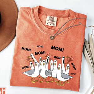 K&ouml;nnte beinhalten: Orangefarbenes Comfort Colors T-Shirt mit einer Grafik von Cartoon-M&ouml;wen, die "MOM!" in schwarzem Text schreien. Das Shirt hat einen Rundhalsausschnitt und kurze &Auml;rmel. Eine silberne Halskette mit Anh&auml;nger liegt auf dem Shirt.
