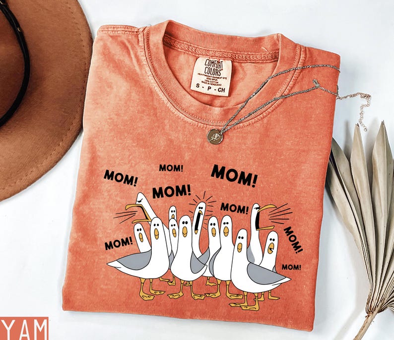 K&ouml;nnte beinhalten: Orangefarbenes Comfort Colors T-Shirt mit einer Grafik von Cartoon-M&ouml;wen, die "MOM!" in schwarzem Text schreien. Das Shirt hat einen Rundhalsausschnitt und kurze &Auml;rmel. Eine silberne Halskette mit Anh&auml;nger liegt auf dem Shirt.