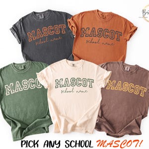 Puede incluir: Cinco camisetas de manga corta en gris carb&oacute;n, &oacute;xido, verde oliva, crema y marr&oacute;n. Cada camiseta presenta la palabra MASCOT en una fuente universitaria con el texto school name en cursiva debajo. El texto inferior dice PICK ANY SCHOOL MASCOT!