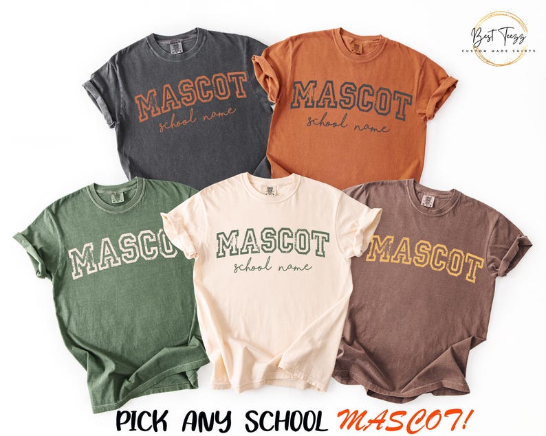 Puede incluir: Cinco camisetas de manga corta en gris carb&oacute;n, &oacute;xido, verde oliva, crema y marr&oacute;n. Cada camiseta presenta la palabra MASCOT en una fuente universitaria con el texto school name en cursiva debajo. El texto inferior dice PICK ANY SCHOOL MASCOT!