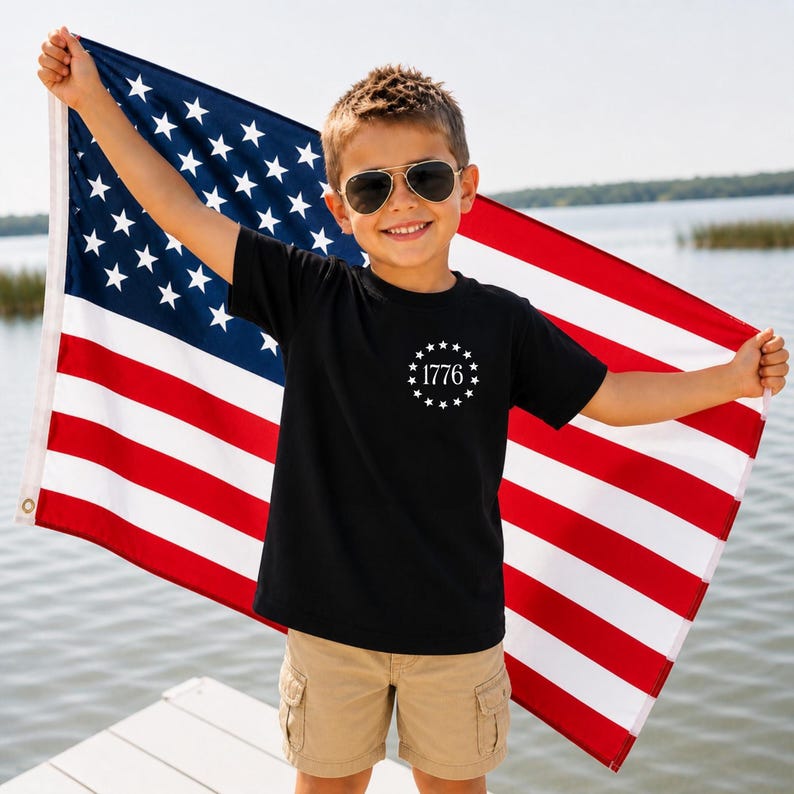 May include: A black short-sleeved t-shirt featuring a white circular graphic on the chest with the text 1776 surrounded by thirteen stars. The shirt is worn with khaki cargo shorts and aviator sunglasses, held against a large American flag.