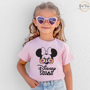 Puede incluir: Camiseta rosa claro con un gr&aacute;fico de Minnie Mouse con gafas de sol te&ntilde;idas y el texto "Disney Squad". La camiseta la lleva una ni&ntilde;a peque&ntilde;a que lleva gafas de sol en forma de coraz&oacute;n.