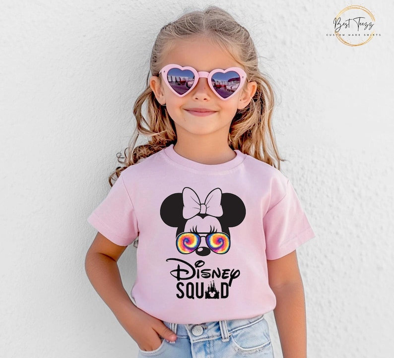 Puede incluir: Camiseta rosa claro con un gr&aacute;fico de Minnie Mouse con gafas de sol te&ntilde;idas y el texto "Disney Squad". La camiseta la lleva una ni&ntilde;a peque&ntilde;a que lleva gafas de sol en forma de coraz&oacute;n.