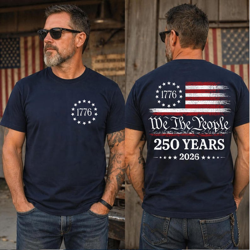 May include: A navy blue short-sleeve t-shirt featuring a white circular star design with the text 1776 on the front. The back displays a distressed American flag, the text We The People, 250 YEARS, and the year 2026 in white lettering.