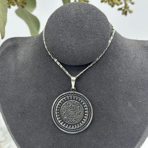 Puede incluir: Un collar de plata con un colgante redondo que presenta un diseño detallado del calendario azteca.