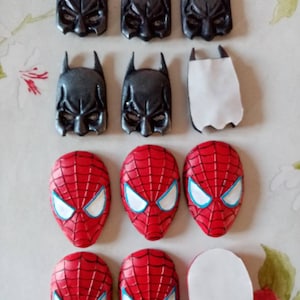 May include: A collection of superhero masks, including Batman and Spiderman, made of black and red fondant. The masks are arranged in rows on a white surface.