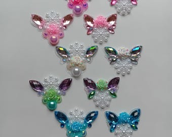 May include: A collection of decorative butterfly embellishments in various colors. Each butterfly features gem-like wings, a central flower, and pearl accents. Colors include pink, purple, blue, and green. The butterflies are arranged on a white surface.