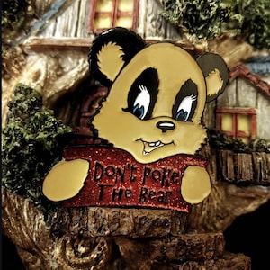 May include: A whimsical bear figurine with a tan face, black ears, and blue eyes. The bear holds a red sign that reads "Don't Poke The Bear." The figurine is set against a backdrop of a miniature house.
