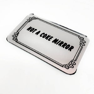May include: A silver rectangular mirror with a black decorative frame and the text "NOT A COKE MIRROR" printed in black.