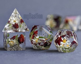 May include: Four clear resin dice with pressed flowers and red berries inside. The dice are in the shape of a pyramid and a cube. The dice are on a gray background.