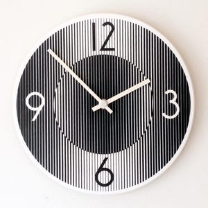 Könnte beinhalten: Eine runde Wanduhr mit einem schwarz-weißen vertikalen Streifenmuster, das eine optische Täuschung einer zentralen Kugel erzeugt. Das Zifferblatt zeigt weiße arabische Ziffern auf den Positionen 12, 3, 6 und 9 Uhr mit dünnen weißen Zeigern.