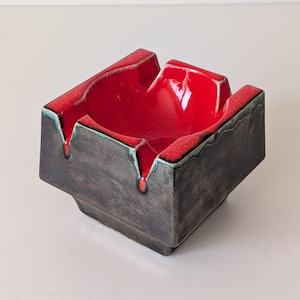 May include: A square ceramic ashtray featuring a deep, glossy red interior basin and a dark, textured charcoal grey exterior. The piece has four distinct notches along the rim, with the red glaze dripping slightly over the dark edges.