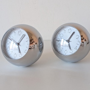 May include: Two spherical table clocks with polished silver-tone metal casings. Each clock features a white circular face with black Arabic numerals from one to twelve, black hour and minute hands, a thin second hand, and the word Quartz printed.