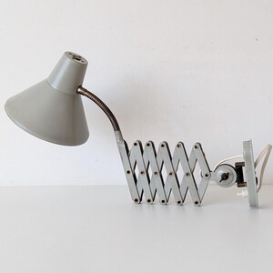 May include: A light grey wall-mounted lamp featuring a conical shade, a flexible metallic neck, and an extendable accordion-style scissor arm mechanism. The fixture includes a circular mounting plate with a white power cord and a small toggle switch.