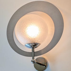 May include: A wall-mounted lamp featuring a circular brushed metal frame surrounding a spherical, textured glass shade. The light emits a warm glow from within the frosted, bubbled glass globe, supported by a polished chrome arm and base.