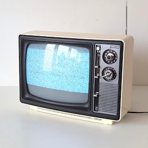 May include: A vintage cream-colored television set with a black front panel, featuring a screen displaying static noise, two circular tuning knobs, a vertical volume slider, and a thin metal antenna extending from the top right corner of the casing.