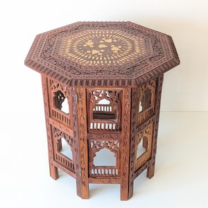 May include: An octagonal wooden side table featuring intricate hand-carved patterns on the sides and legs. The tabletop displays a central inlaid floral design in a lighter wood tone, surrounded by a detailed carved border and open archway panels.