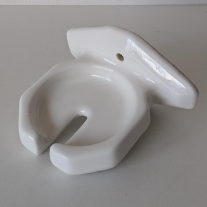 May include: A white ceramic wall-mounted soap dish with a unique, curved design. The dish features a central circular area with a slot and a raised, angled section for mounting. The glossy finish reflects light, highlighting its smooth, clean lines.