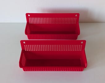 May include: Two bright red wall-mounted storage baskets. Each basket has a rectangular shape with a perforated top section and a solid base. The baskets are designed with a small hole for hanging. Ideal for organizing small items.