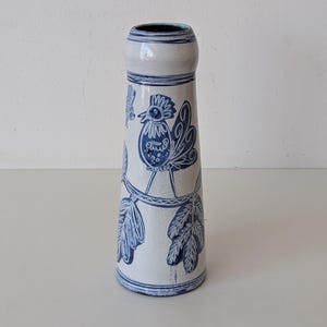 Può includere: Un vaso in ceramica bianca con disegni di gallo e foglie blu dipinti a mano. Il vaso ha una forma cilindrica con base svasata e collo stretto. Il gallo è raffigurato con dettagli intricati e le foglie aggiungono un elemento naturale.