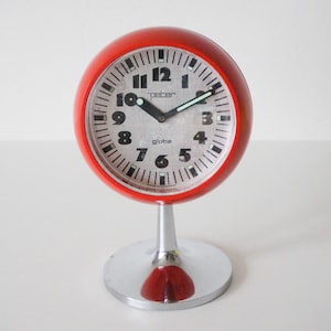 May include: A red, spherical "Peter Globe" alarm clock with a white face and black numerals. The clock has a silver base and a red, teardrop-shaped accent. The hands are black with a green second hand.