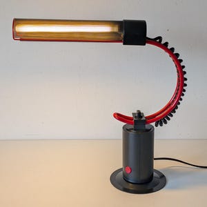 May include: A black and red desk lamp with a curved arm and a cylindrical base. The lamp has a red accent on the arm and a red button on the base. The lamp's light is encased in a rectangular shade.