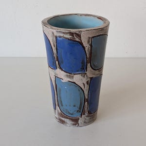 Könnte beinhalten: Eine zylindrische Keramikvase mit einem Muster aus blauen Formen auf beigem Hintergrund. Das Innere der Vase ist hellblau. Die Vase wird wahrscheinlich zu dekorativen Zwecken verwendet. Die Vase ist etwa 20 cm hoch.
