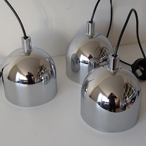 May include: Three chrome pendant lights with black cords and a black ceiling mount. The lights have a reflective, rounded dome shape. The lights are on a white surface.