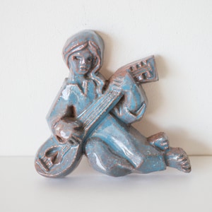 May include: A blue ceramic wall sculpture of a person playing a stringed instrument. The sculpture has a rustic, vintage look.