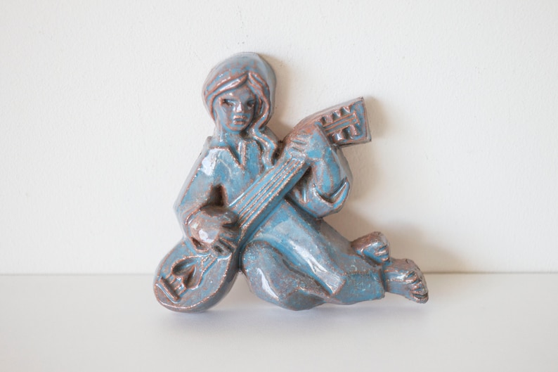 May include: A blue ceramic wall sculpture of a person playing a stringed instrument. The sculpture has a rustic, vintage look.
