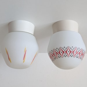 May include: Two flush mount ceiling light fixtures with white frosted glass globes. The left globe features yellow and red brushstroke accents. The right globe displays a repeating red diamond pattern outlined in black lines and dots.