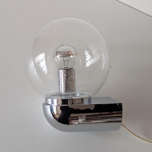 May include: A vintage wall-mounted light fixture with a clear, speckled glass globe. The light bulb is visible inside the globe. The fixture has a chrome-colored, rectangular base and a curved, polished metal support.