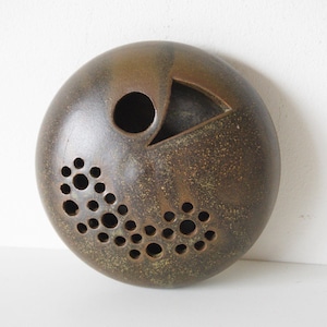 May include: A circular, bronze-toned ceramic wall decoration with a textured surface. It has a large circular aperture, a triangular cut-out, and a cluster of smaller holes, forming a distinctive decorative pattern. The piece has a speckled finish.