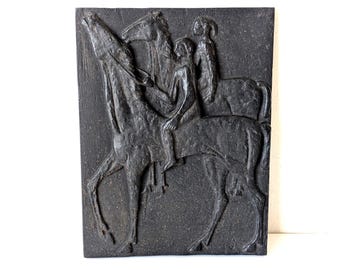 May include: A rectangular black wall plaque featuring a raised relief sculpture of two horses and two riders. The dark, textured surface depicts the figures in profile, with one horse rearing its head upward while the riders sit atop the animals.