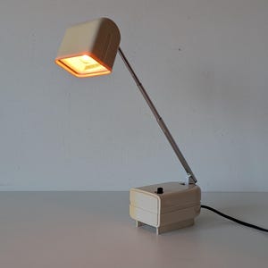 May include: A vintage desk lamp with a cream-colored rectangular base and a matching square lampshade. The lampshade is angled and connected to the base by a silver-colored adjustable arm. The lamp is plugged in and lit, emitting a warm orange glow.