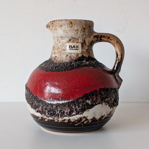 May include: A ceramic pitcher with a textured surface and a handle. The pitcher features a color scheme of red, black, white, and brown. The label on the pitcher reads "BAY. KERAMIK".