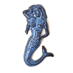 Op de afbeelding: Een decoratief wandfiguur in de vorm van een zeemeermin, gemaakt van keramiek met een glanzend, gespikkeld donkerblauw en zwart glazuur. De zeemeermin staat in een verticale houding met één arm achter het hoofd en een geschubde staart.