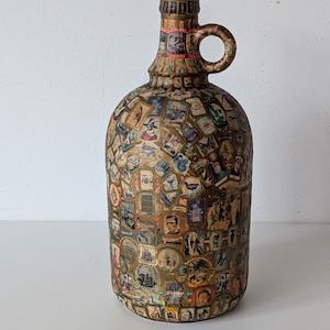 Può includere: Una brocca decorativa in stile vintage, ricoperta da un collage di francobolli e immagini colorate. La brocca ha un corpo arrotondato e un collo stretto. I francobolli raffigurano varie scene e ritratti, con una combinazione di colori marrone e oro.