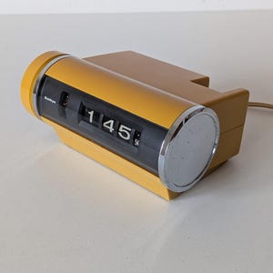 Könnte beinhalten: Ein senfgelber Klappwecker, der die Uhrzeit 1:45 anzeigt. Die Uhr hat ein zylindrisches Zifferblatt mit einem verchromten Ende und einem rechteckigen Sockel. Ein Netzkabel ist sichtbar.