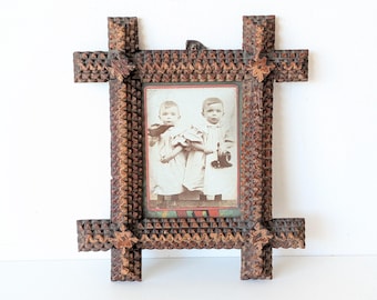 May include: A rectangular wooden picture frame featuring intricate chip-carved geometric patterns and extended corner joints. The frame holds a vintage black and white photograph of two young children wearing light-colored dresses and holding small objects.