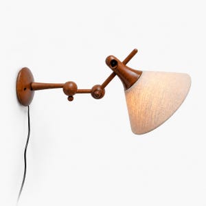 Könnte beinhalten: Eine Wandlampe aus Holz mit einem hellbeigen Stoffschirm. Die Lampe verfügt über eine Reihe von Holzgelenken, die eine verstellbare Positionierung ermöglichen. Sie wird mit einer runden Holzbasis und einem schwarzen Netzkabel an der Wand befestigt.