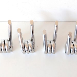 Puede incluir: Un juego de cuatro ganchos de pared de metal plateado, cada uno con una base en espiral y dos puntas orientadas hacia arriba. Los ganchos tienen un acabado liso y reflectante, con una forma que recuerda a un gesto de la mano.