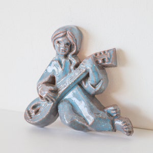 May include: A blue ceramic wall sculpture of a person playing a guitar. The sculpture is in a sitting position with the guitar resting on their lap.