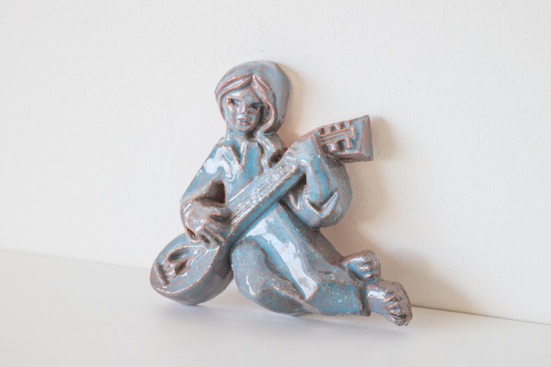 May include: A blue ceramic wall sculpture of a person playing a guitar. The sculpture is in a sitting position with the guitar resting on their lap.
