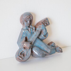 May include: A blue ceramic wall sculpture of a person playing a guitar. The sculpture is in a seated position with the guitar resting on their lap.