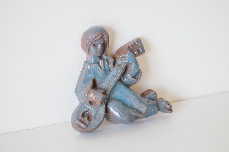 May include: A blue ceramic wall sculpture of a person playing a guitar. The sculpture is in a seated position with the guitar resting on their lap.