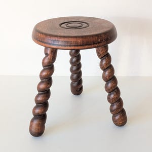May include: A dark brown wooden stool featuring a circular seat with a carved concentric circle design in the centre. The stool is supported by three legs, each crafted with a distinct twisted, spiral rope-like pattern throughout their length.