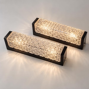 May include: Two rectangular wall sconces featuring black metal end caps and a textured, clear glass shade with a geometric, faceted pattern. The lights are illuminated, casting a warm glow and creating a starburst effect on the surface below.