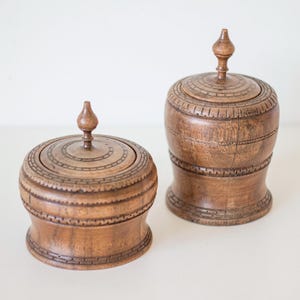 Könnte beinhalten: Zwei braune Holzbehälter mit Deckel und geschnitzten Mustern. Die Behälter haben eine runde Form und eine dekorative Spitze oben. Der kleinere Behälter ist breiter und kürzer als der größere. Die Behälter sind aus Holz.