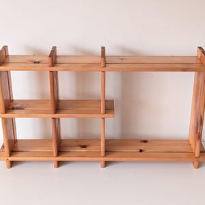May include: A light brown wooden wall shelf featuring a modular design with three distinct sections. The unit is constructed from horizontal planks and vertical dividers, creating a tiered storage structure with visible wood grain and knots.